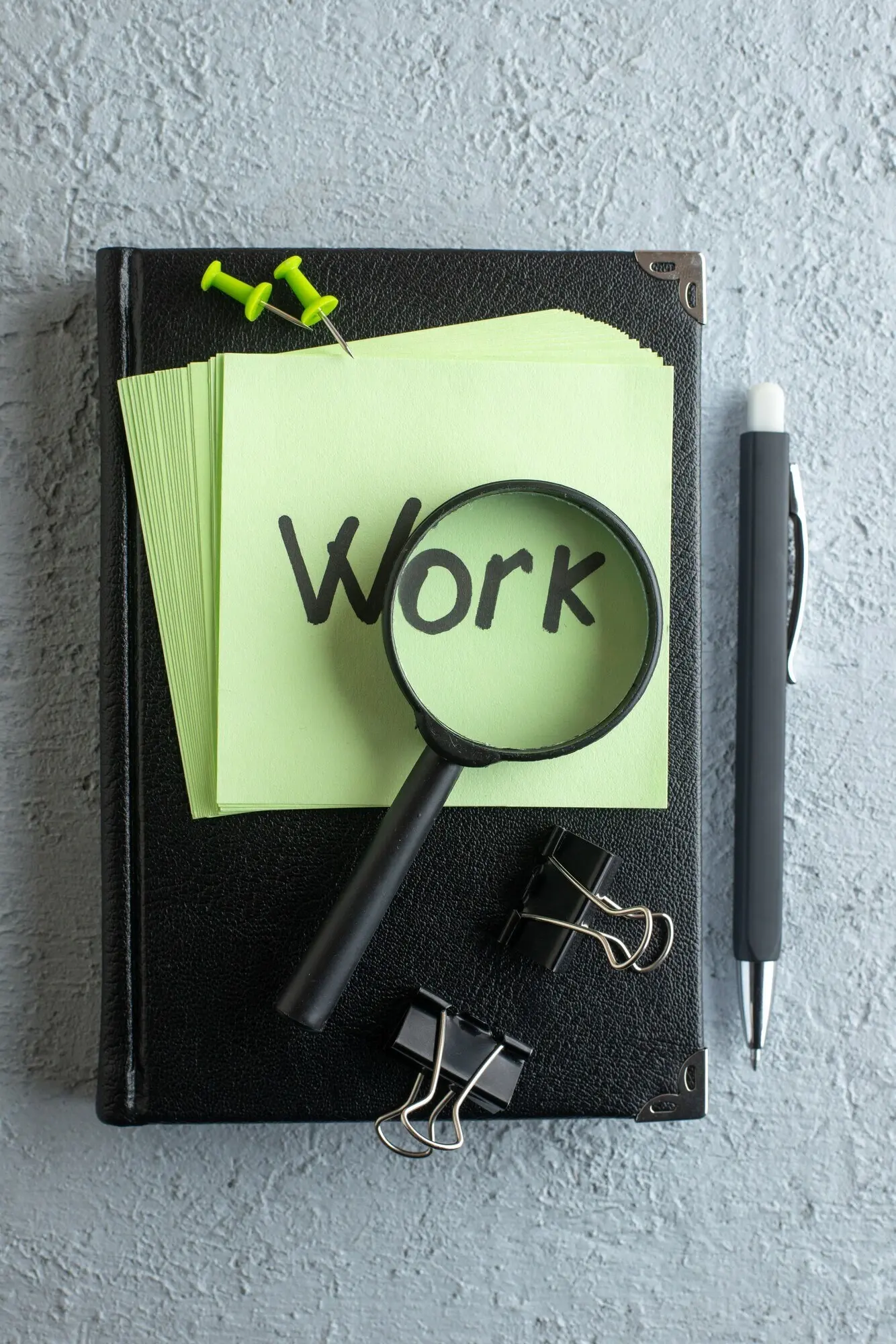 Draufsicht: geschriebene Arbeitsnotiz auf grünen Stickern mit Notizblock und Lupe auf weißem Hintergrund; College, Farbe, Job, Schule, Büro, Business, Schreibhefte.